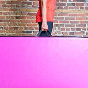 Female athlete holding a pink PRISP gymnastics mat by its handles, in front of brick wall